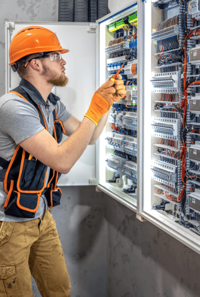 A man wearing an orange safety vest and glasses is focused on repairing a control panel.